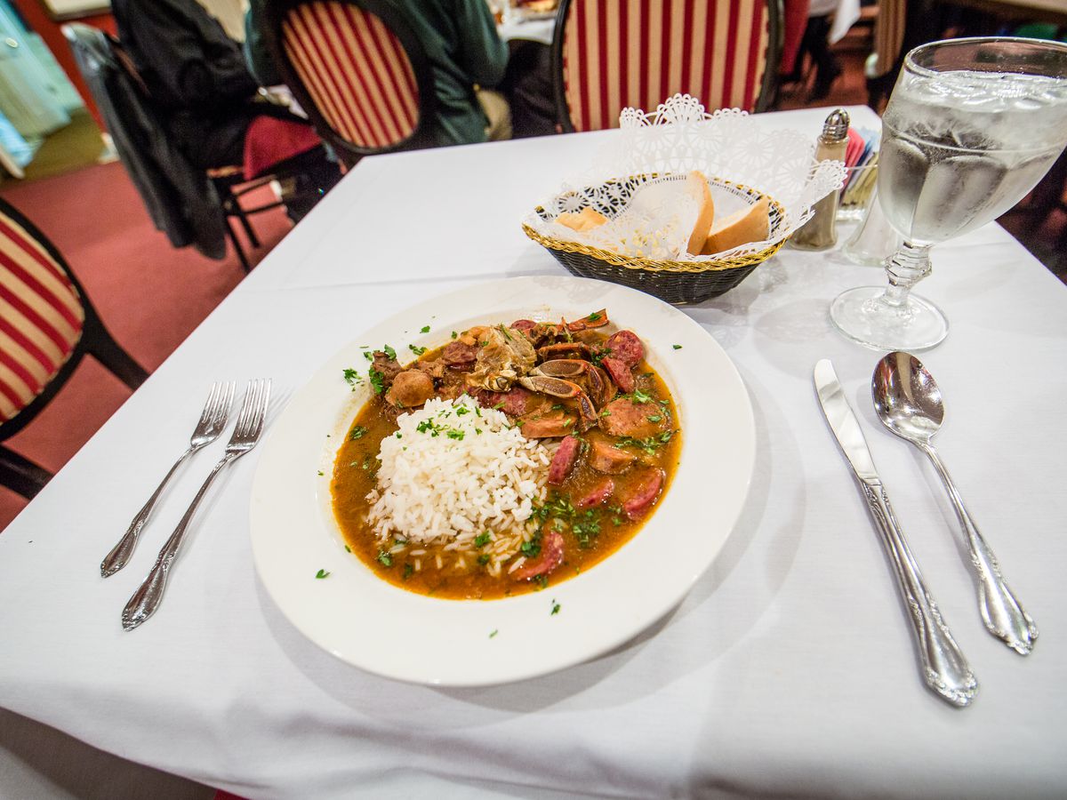 A view of a bowl of Creole gumbo and a bread basket on a table with a white tablecloth and red striped chairs.