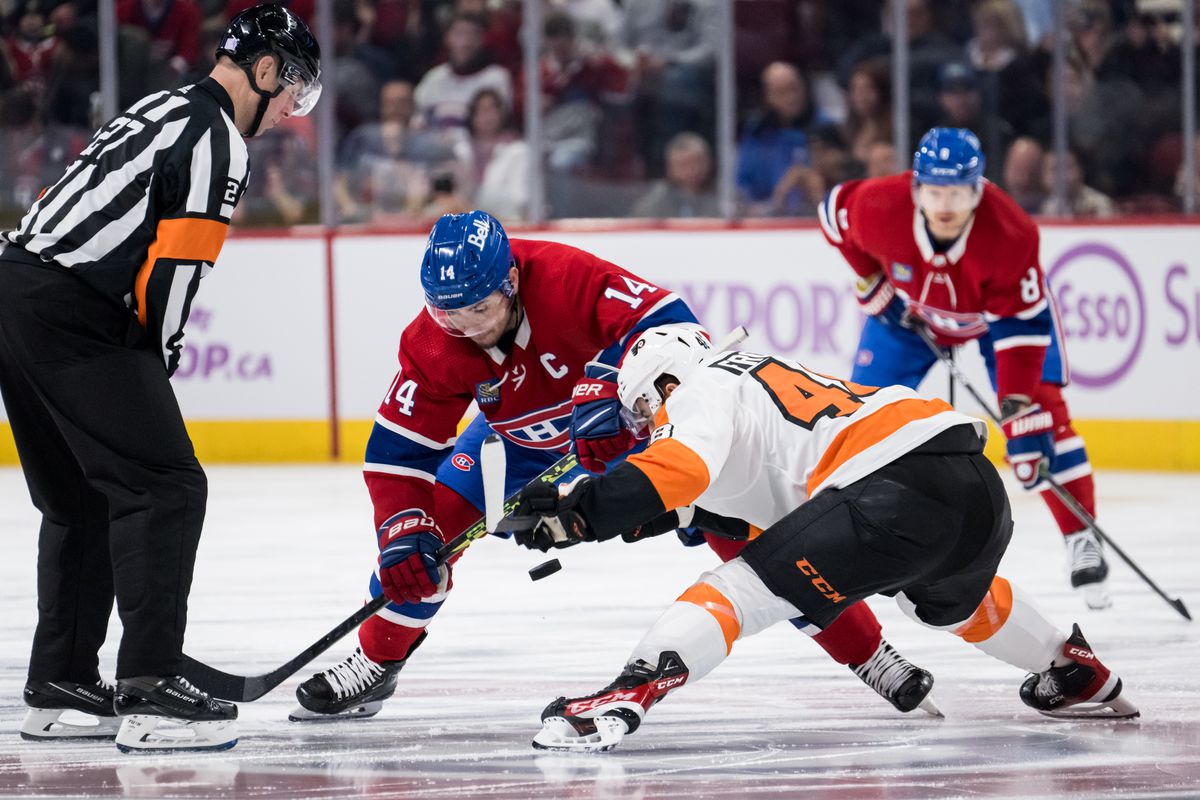 Morgan Frost, with back turned, is dropped down low for a face off against Nick Suzuki. Frost is in the Flyers’ white away jersey and Suzuki is in the Canadiens’ red home jersey. A referee stands to the left after dropping the puck, and in the background is defenseman Mike Matheson