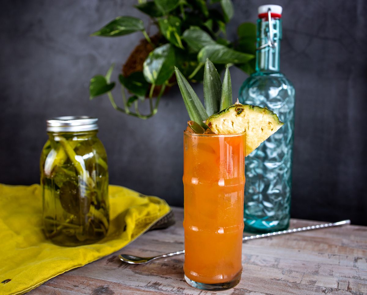 A deep orange drink in a tall glass with a wedge of pineapple on the rim. In the background is a blue glass bottle, stirring spoon, and a jar with green stuff in it.