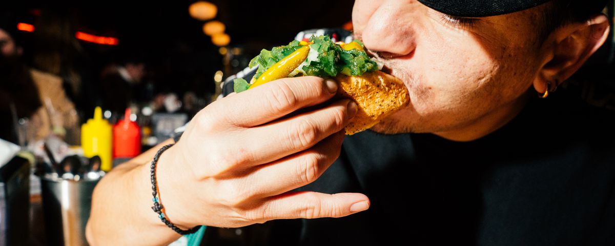An employee at a bar takes a bite of a Chicago-style hot dog with relish and sport peppers.
