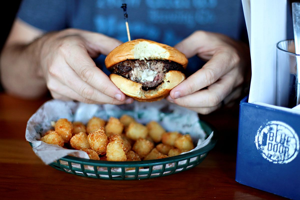A person holding a burger with a bite out of it, cheese is oozing out of the center, there is also a basket of tater tots on the table. 