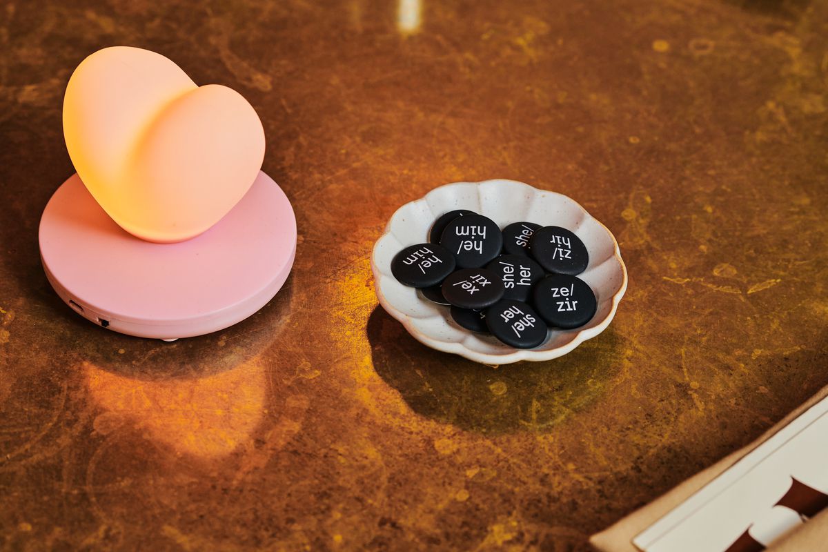 Small bowl containing button pins reading “he/him,” “she/her,” and “ze/zir” next to a lit heart-shaped lamp.