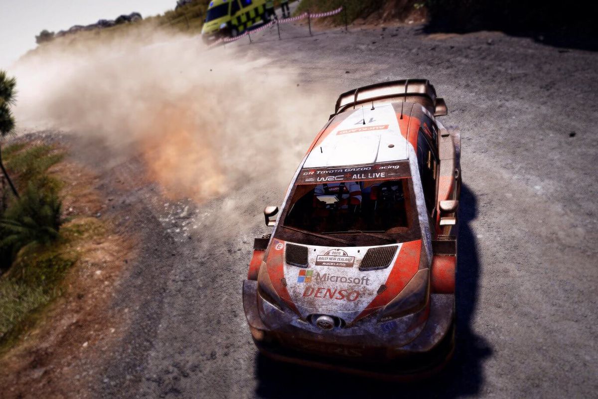 a dirty rally car, trailing dust from a gravel road, slides through a turn