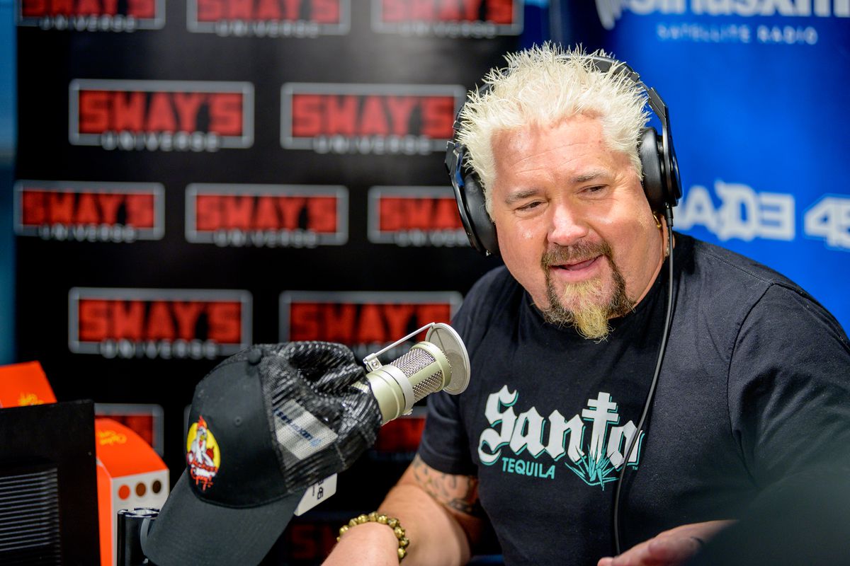 With his signature spiked blond hair and goatee, Guy Fieri wears headphones and a dark blue t-shirt while recording a radio interview.