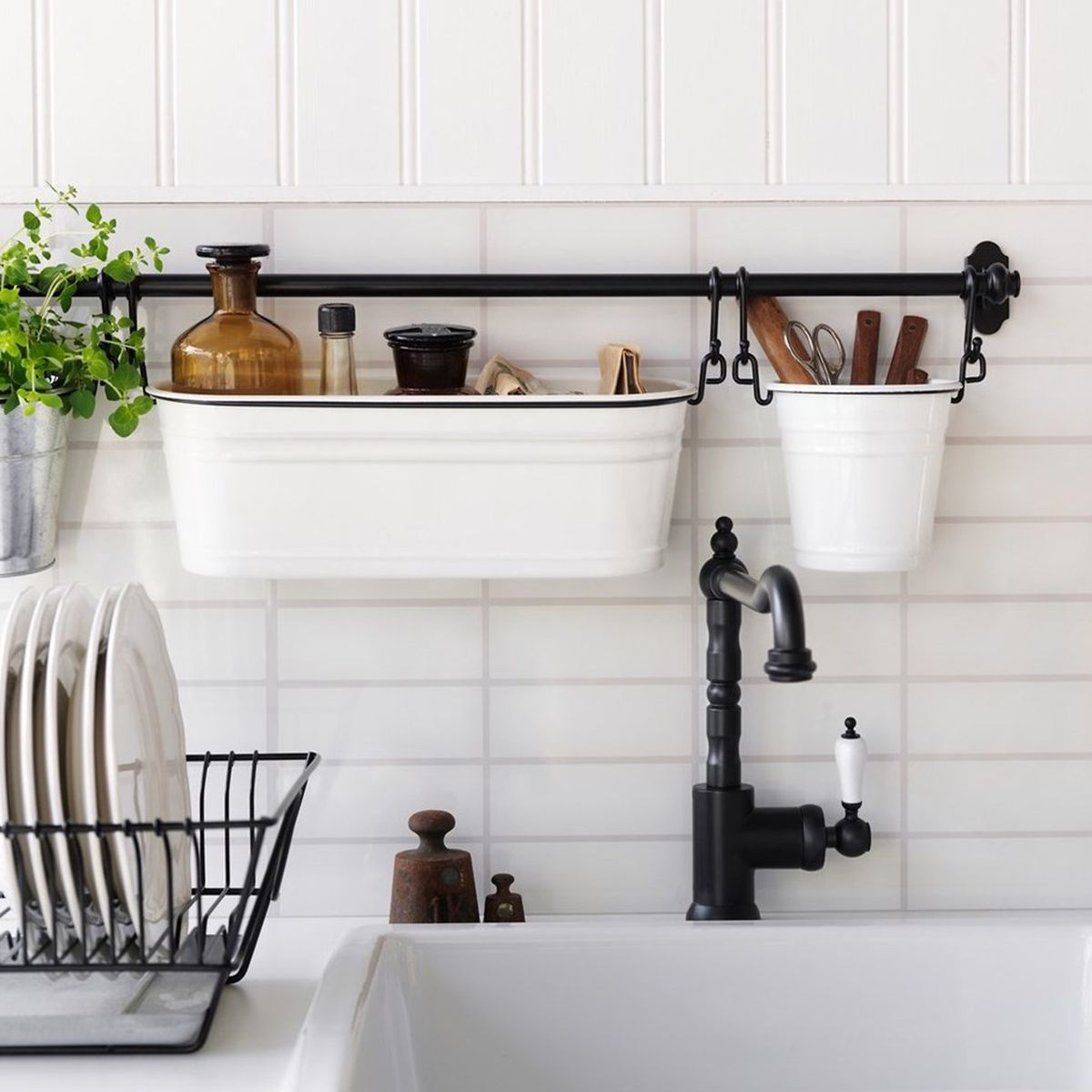 A kitchen has a deep white farmhouse sink, white dishes drying in a black rack, and white buckets hanging from a black bar behind the sink.
