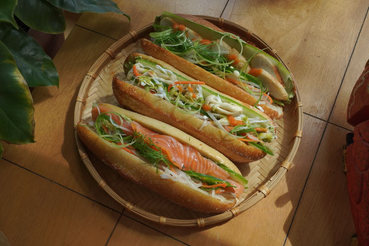 Three banh mi with different fillings sit on a straw tray on a beige tiled floor