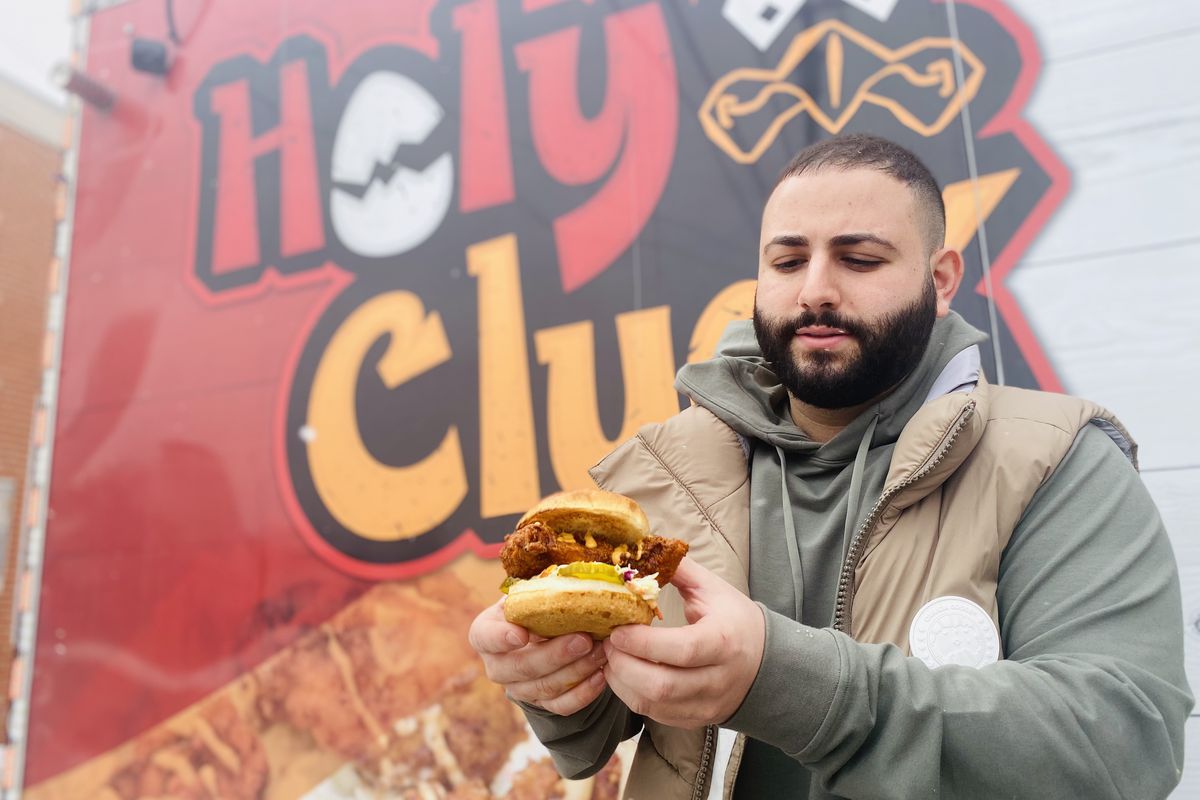 Man wearing a puffy vest and hoodie sweatshirt, holding a spicy chicken sandwich in front of a food truck in Dearborn, Michigan