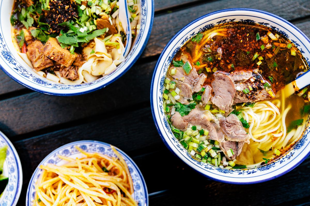 Two blue-rimmed white bowls of beef noodles sit atop a table next to a smaller blue plate of potatoes.