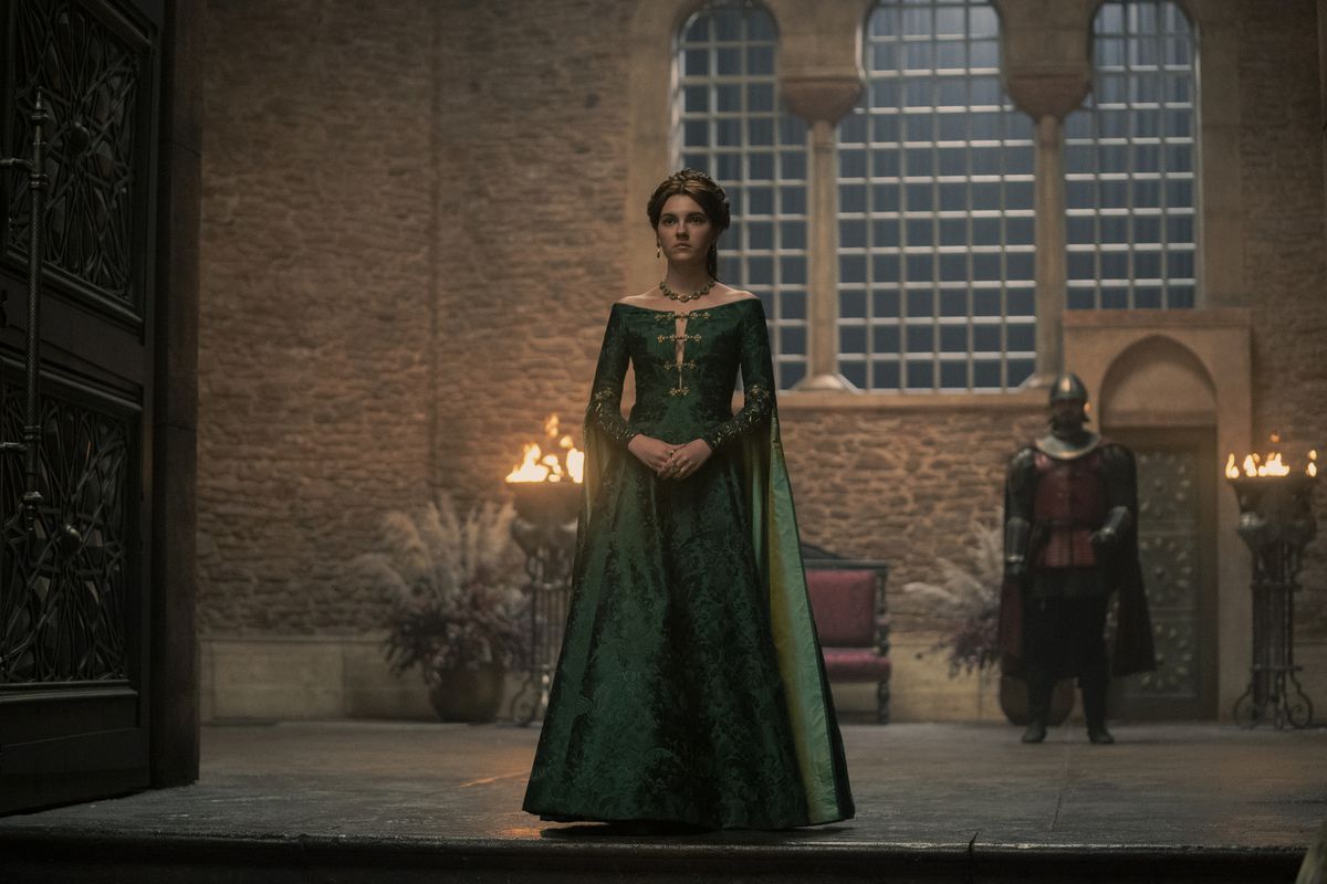 Alicent standing in the doorway to the great hall in a green dres, with her hands clasped in front of her at her waist
