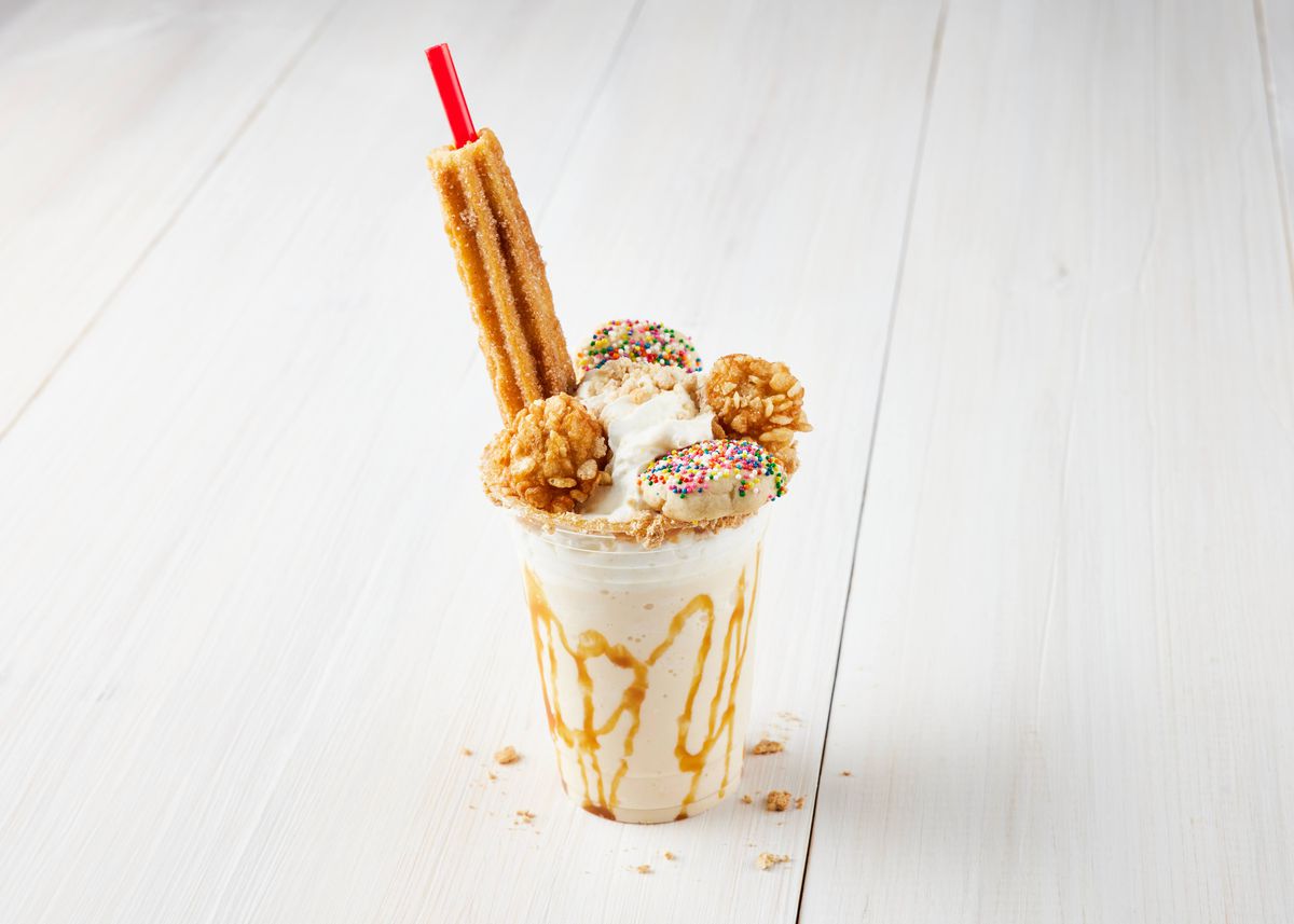 A milkshake sits in a clear plastic glass with a straw inside a churro.  It is topped with jimmies and cookies.