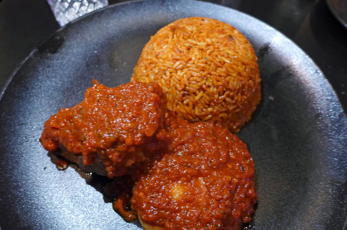 Two pieces of chicken thickly coated with red sauce next to a red ball of rice on a black plate.