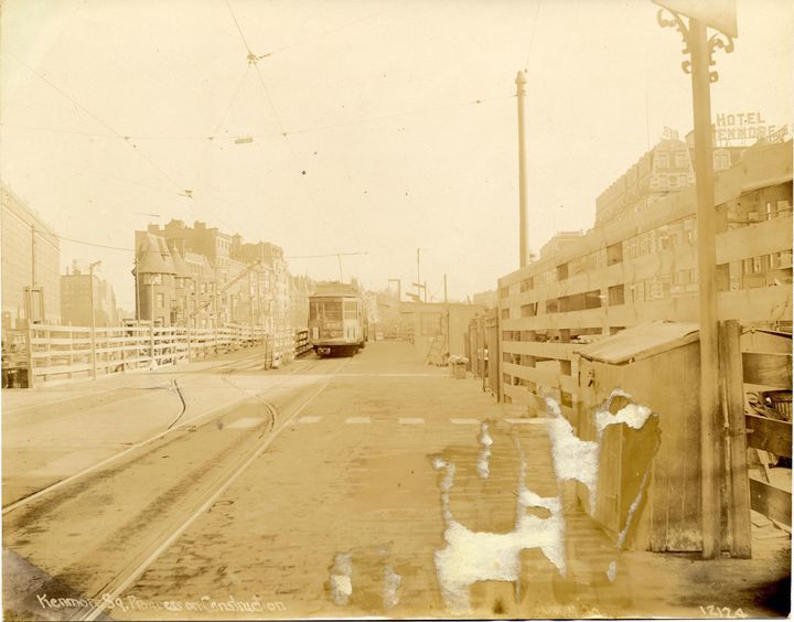 A black-and-white photo of a train trundling down a track in a largely empty urban landscape. 