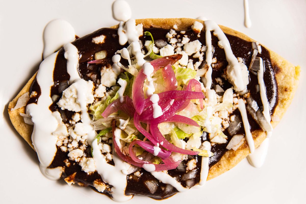 A thick, oval tortilla topped with brown mole, white cheese and sauce, and slices of red onion.
