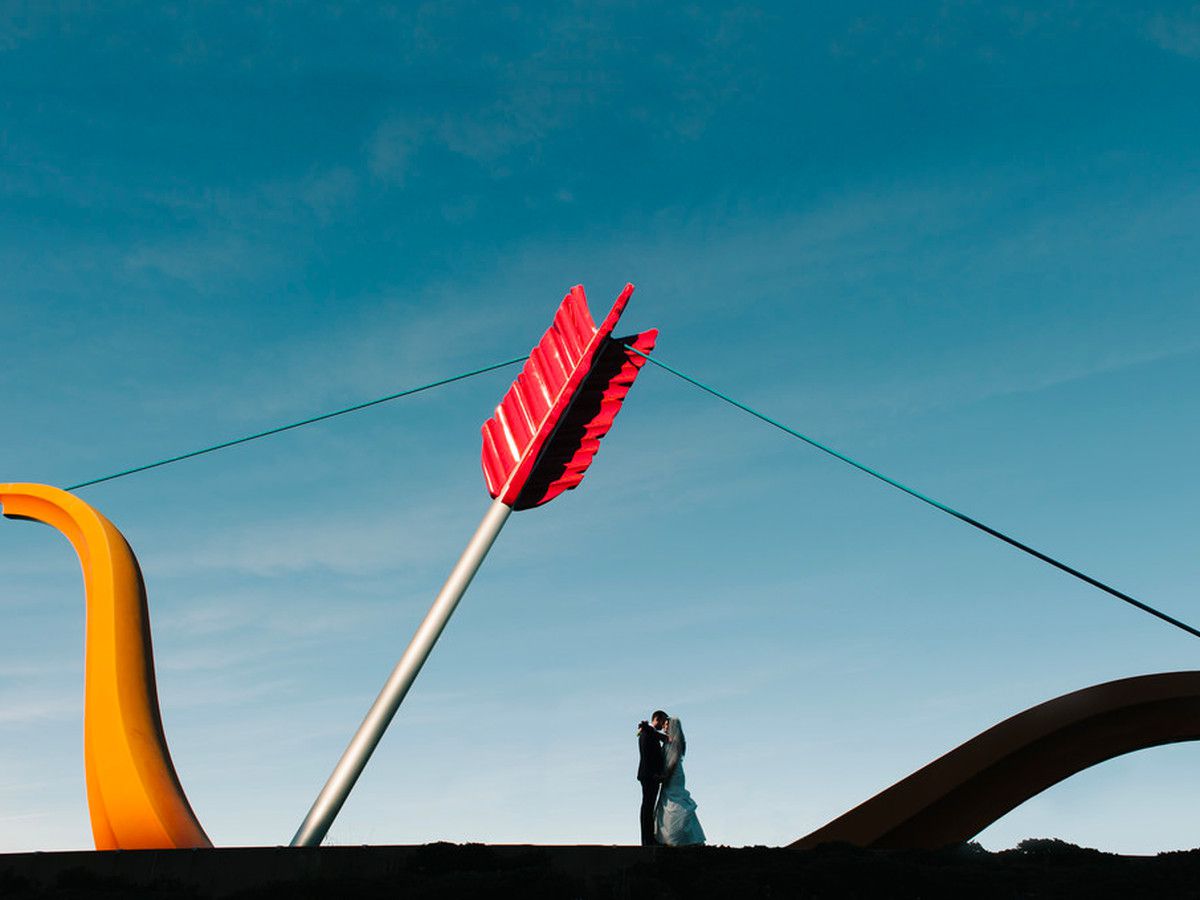 Two people embrace next to Cupid’s Span in San Francisco. Cupid’s Span is a large bow and arrow that rises from the ground. 
