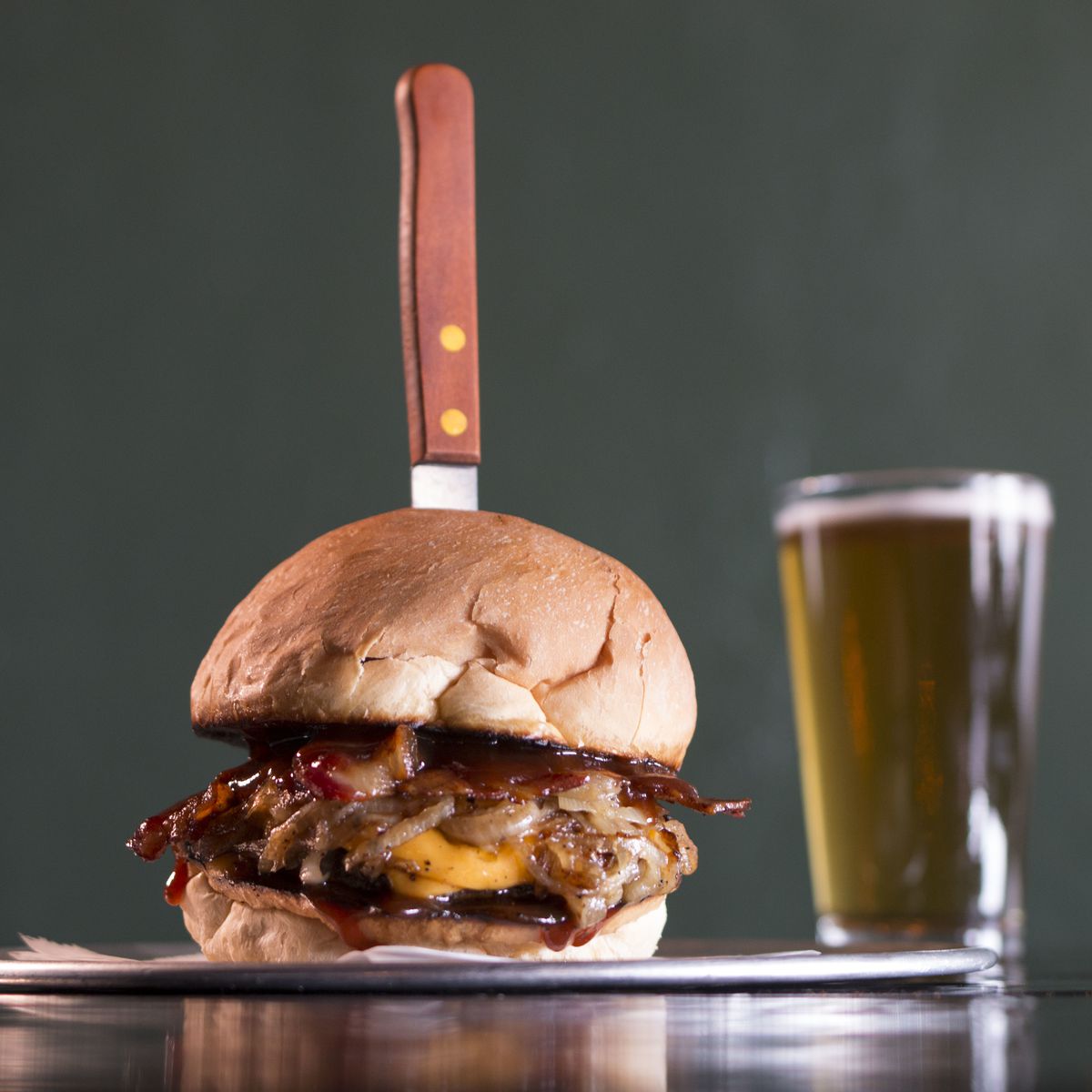 A side view of the BBQ bacon cheeseburger at the Balboa being stabbed with a steak knife. A pint of pale beer sits to the right in the background. 