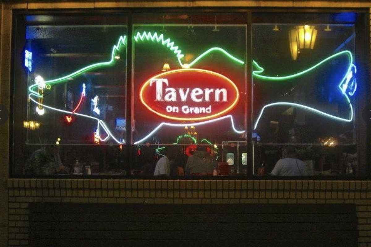 A dark window storefront with a large neon sign in the shape of a fish, with a logo in the middle that says “Tavern on Grand.”