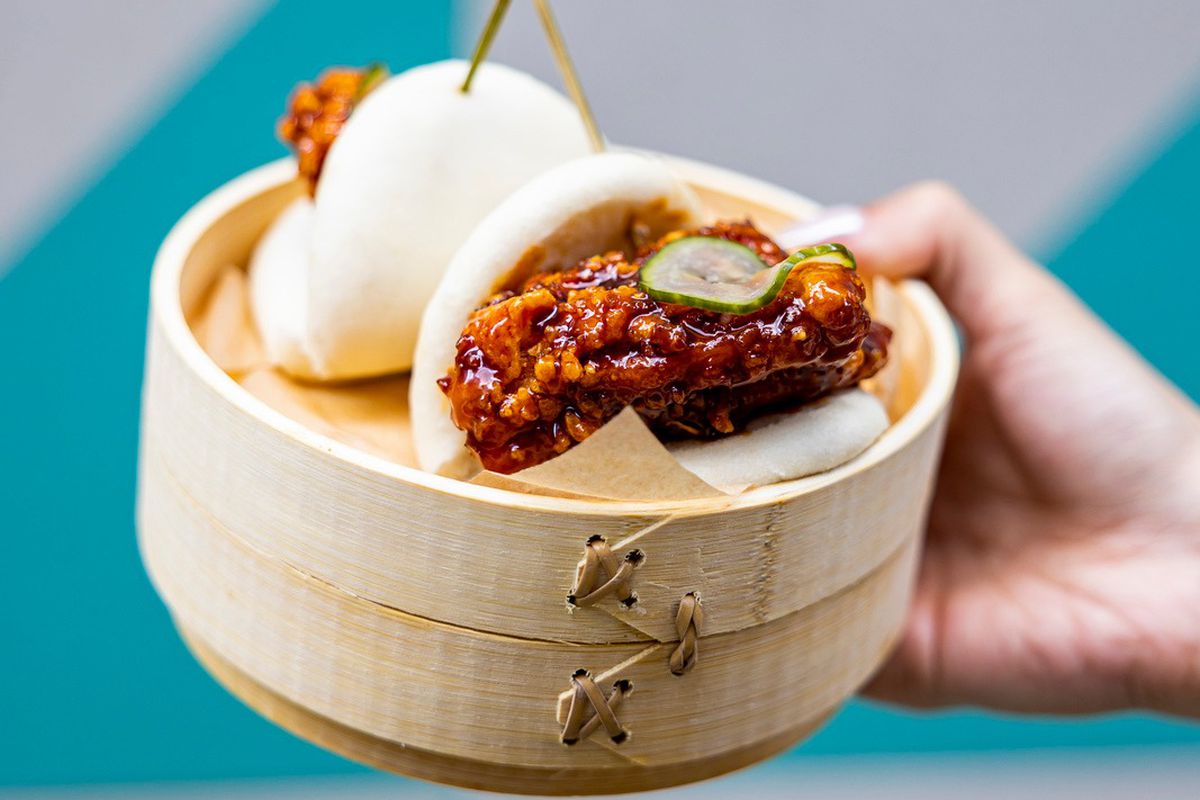 Two Seoul hot chicken bao tossed in garlic gochujang sauce topped with spicy pickles sit in a wooden steamer basket from Hawkers Asian Street Fare.