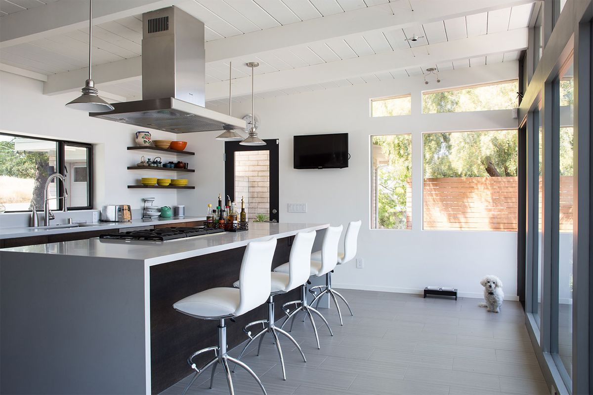 A midcentury modern kitchen with a grey floor and grey kitchen island. The ceiling is wooden and painted white. There are tall windows. There is a small white dog sitting in a corner.