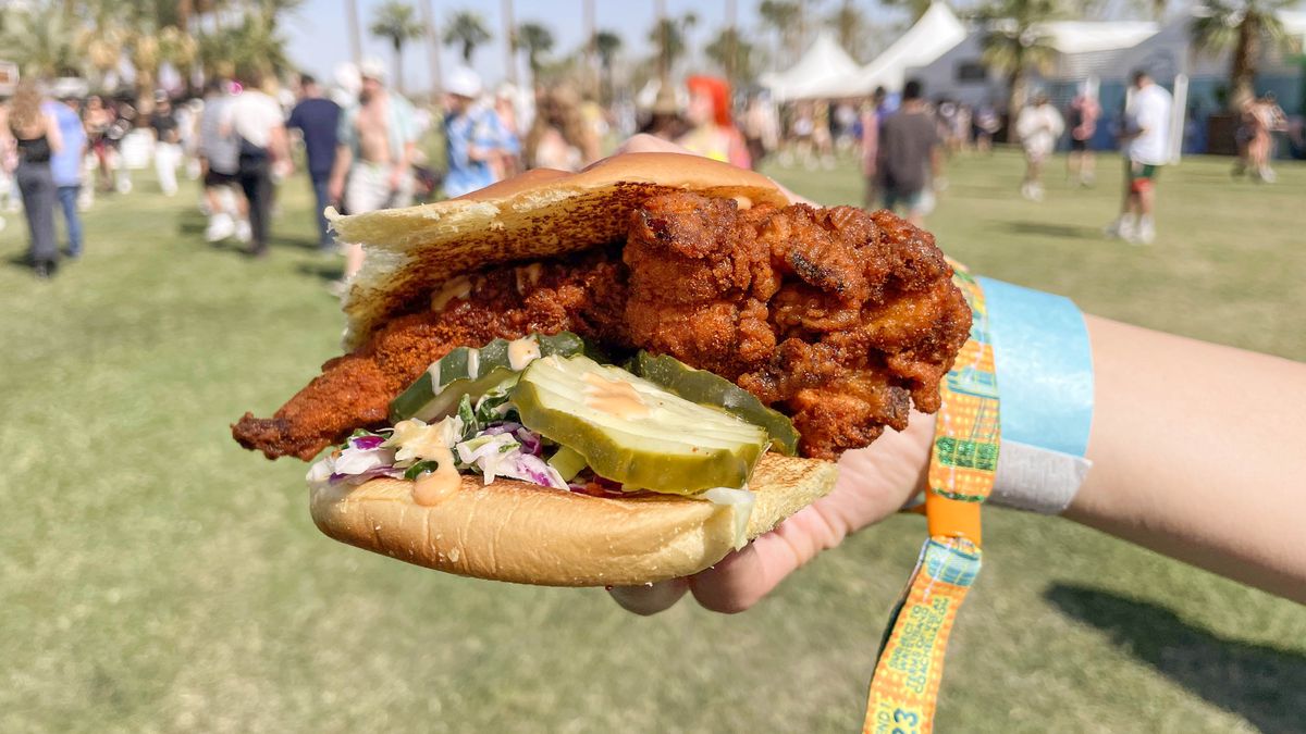 A hand, at right, enters frame holding a fried chicken sandwich with pickles on the grounds of a music festival.
