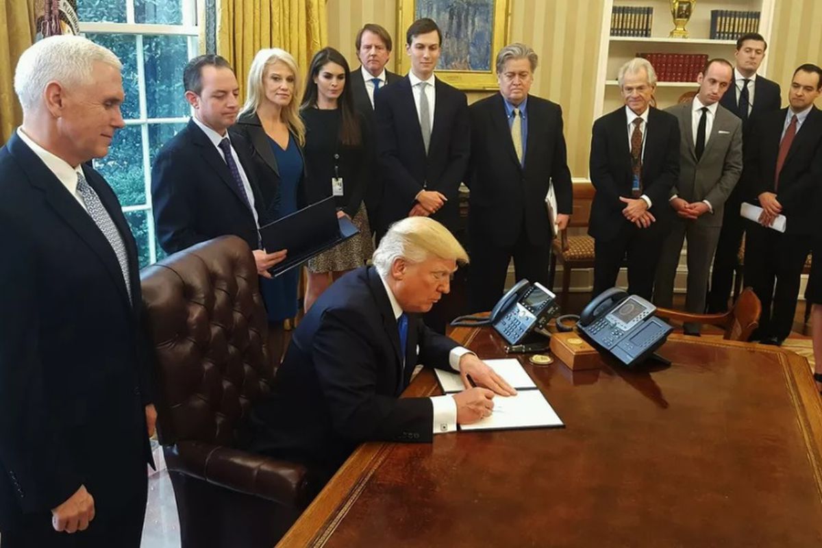 Donald Trump and various cohorts surrounding a desk while he signs an order.