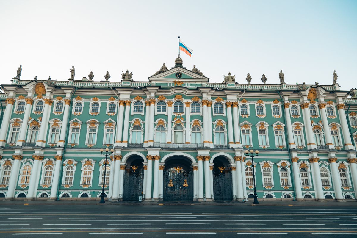 The exterior of the Hermitage Museum in Russia. The facade is green and white. There are columns flanking the entrance. There is a flag on flagpole on the roof above the entrance. 
