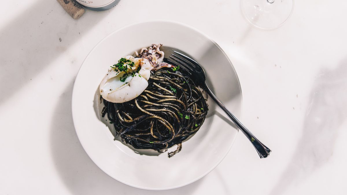 A white bowl with black pasta in it on a white tablecloth. 