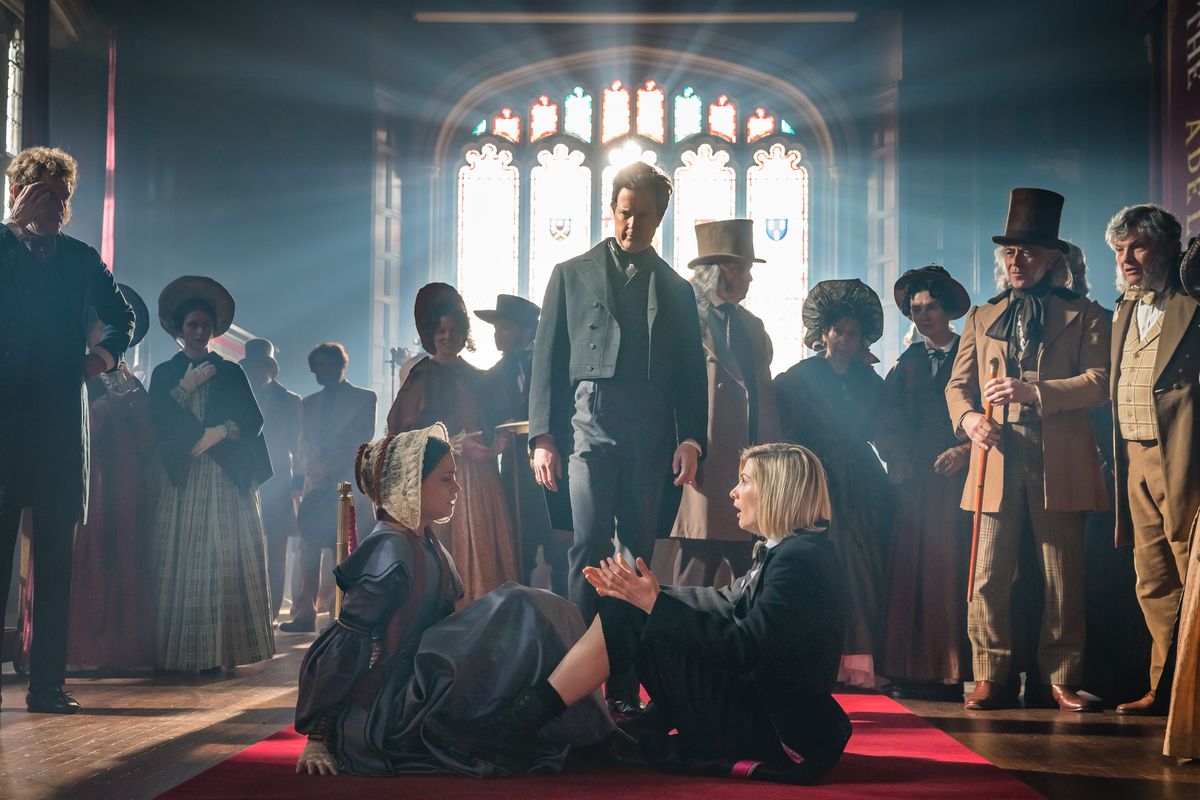 Jodie Whittaker as the Doctor in Doctor Who, sitting on the floor of a room with light streaming in through a stained glass window, surrounded by Ada Lovelace, Charles Babbage and a crowd of curious onlookers.