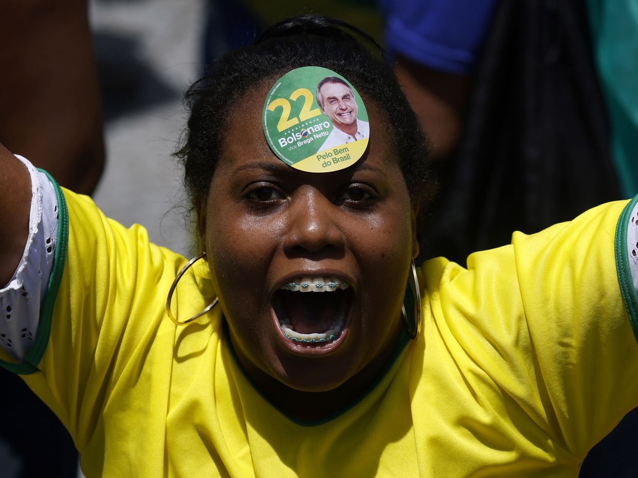 A woman in a yellow shirt wears a sticker on her forehead supporting Bolsonaro in 2022. Her arms are raised and she is cheering. 