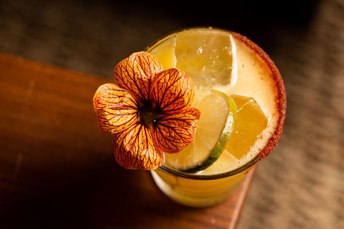 Colorful cocktail with tequila, americano, lime, passionfruit, mango, and smoked cayenne salt with flower garnish.