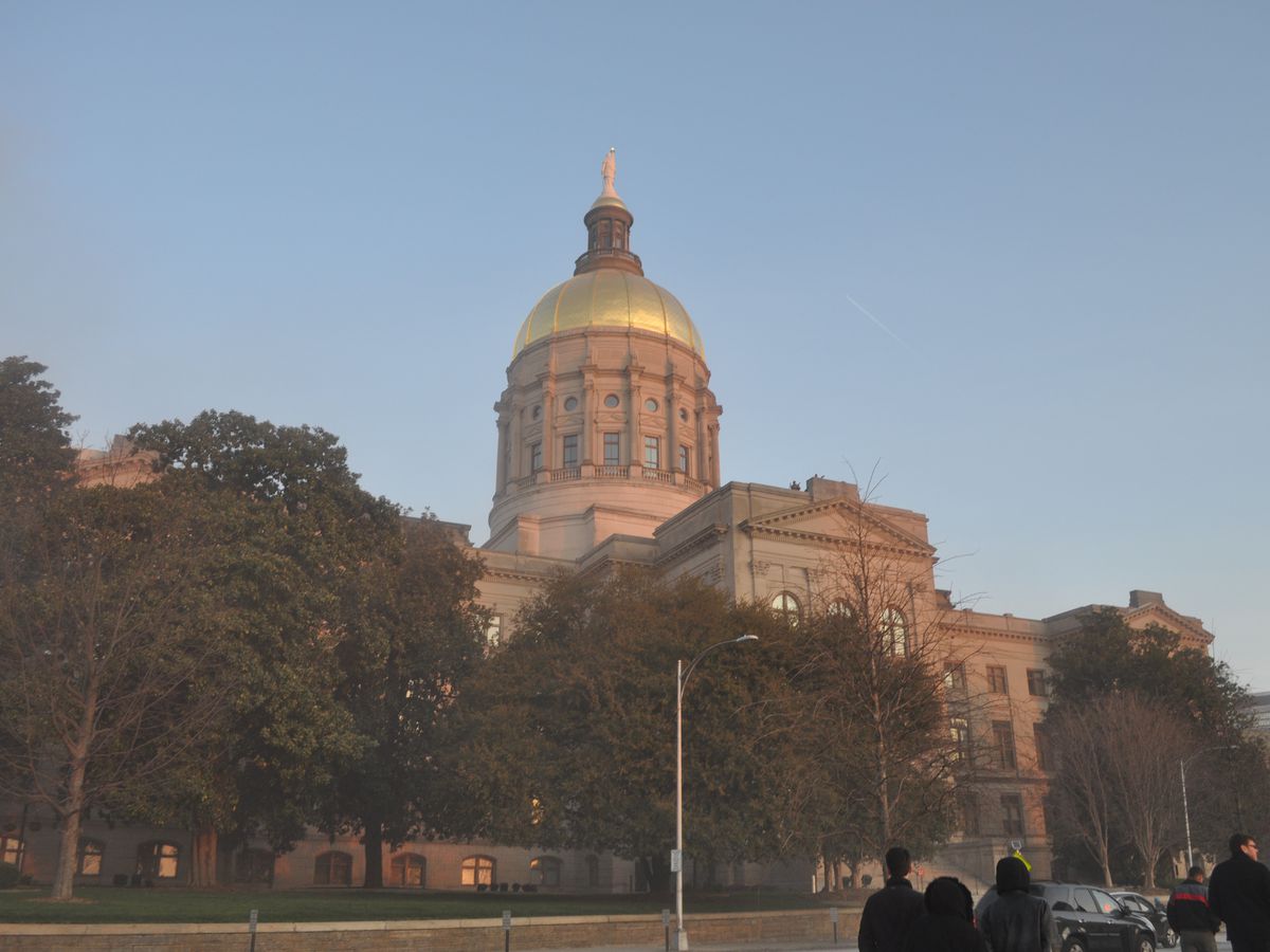 The exterior of the Georgia State Capitol in Atlanta. The building has a tower with a gold dome on top. There are trees in front of the building. 
