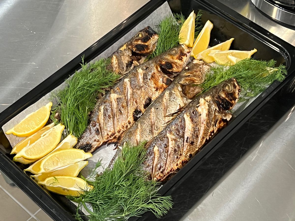 Grilled branzino with herbs and lemon on a black plastic tray.