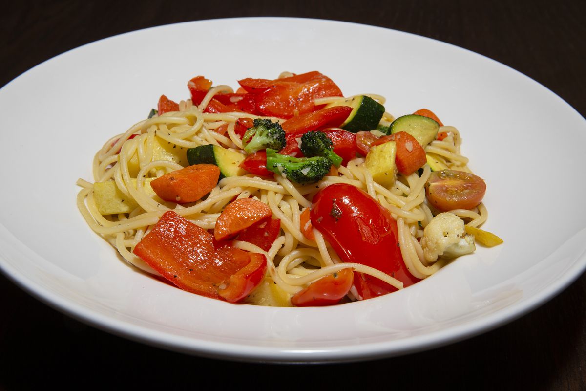 A white bowl filled with cooked spaghetti and colorful roasted vegetables like red peppers and green broccoli and zucchini.