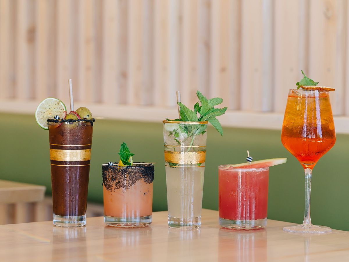 Five colorful cocktails on light wood table with green booth behind them.