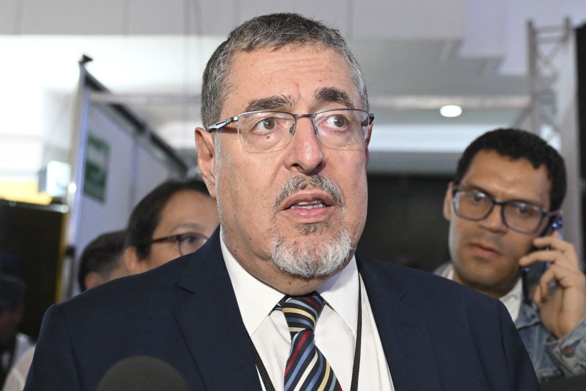 Arévalo, pictured from the shoulders up, wears frameless glasses, a navy suit jacket, and a striped tie. Behind him, a man speaks on a cellphone.