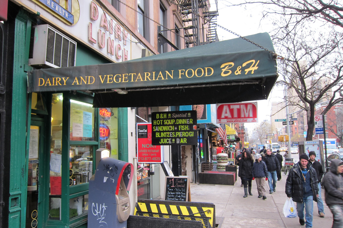 A green awning hangs over a New York City sidewalk busy with pedestrian. In a yellow printed font, it reads “Dairy and Vegetarian Food.”