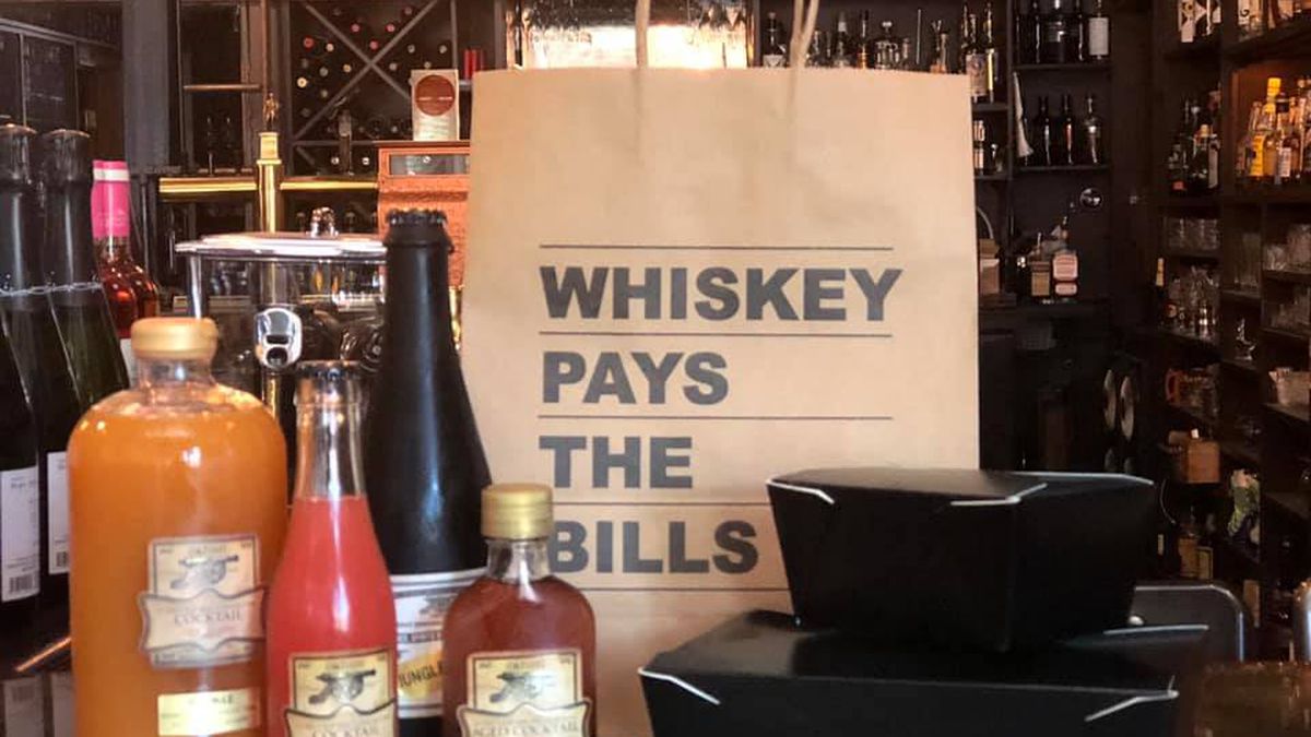 A collection of bottled cocktails at Canon, with a large paper to-go bag that says “Whiskey Pays the Bills”