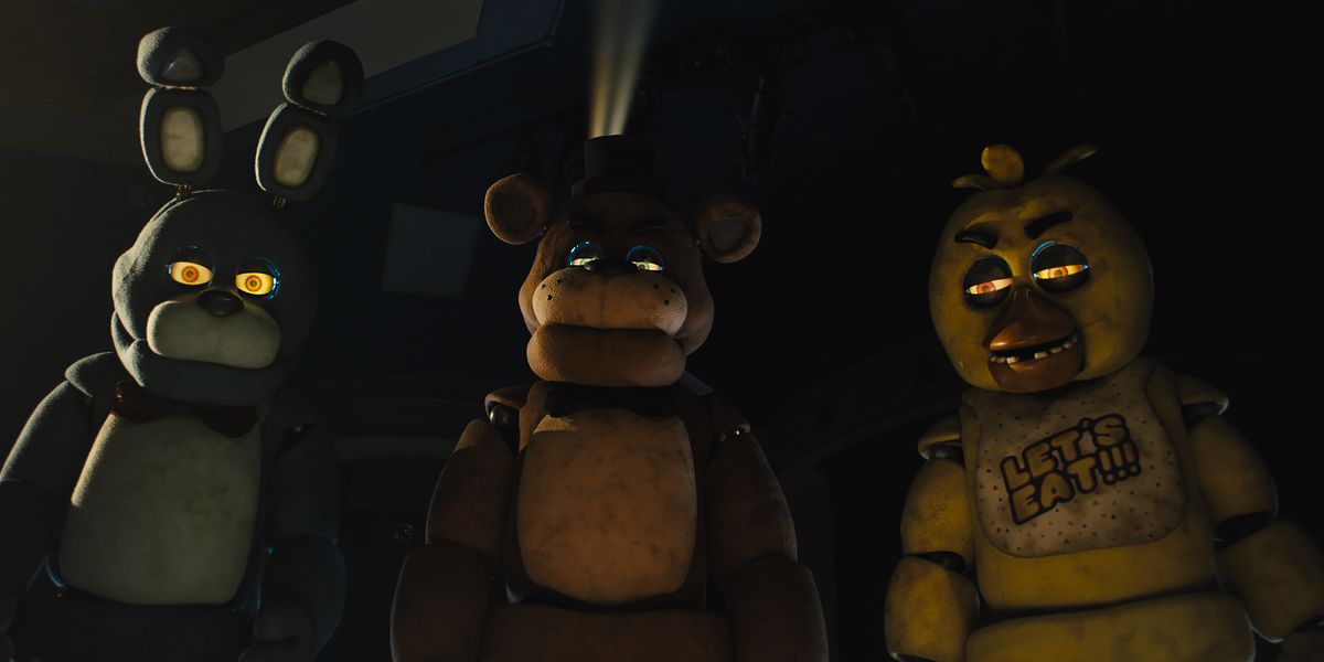 Animatronics villains Bonnie, Freddy Fazbear, and Chica loom in a dark space, narrowed eyes glowing threateningly, in the Five Nights at Freddy’s movie