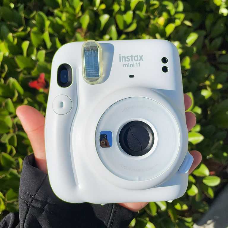 A hand holding Fujifilm's white Instax Mini 11 instant camera in front of a bush outside.