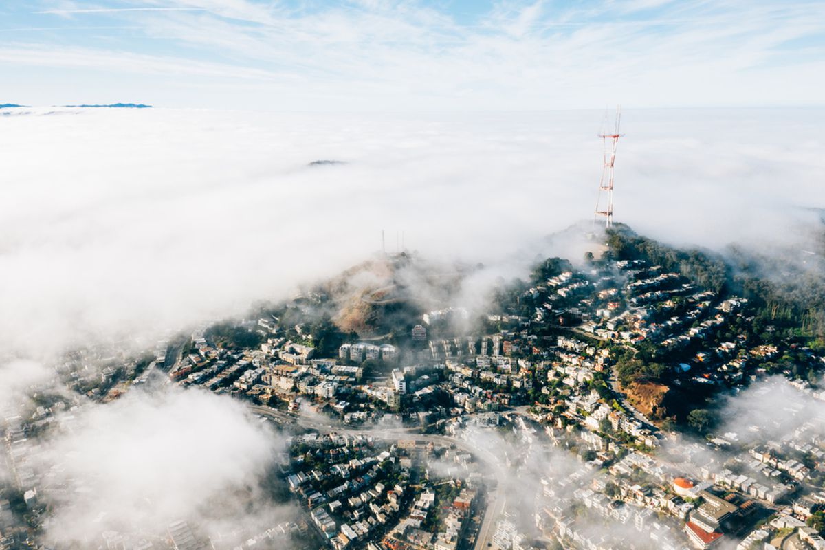 A high aerial photo of homes in the San Francisco hills, with patches of heavy white fog obscuring much of the landscape. A three-pronged broadcast antenna rises hundreds of feet into the air from a hilltop.