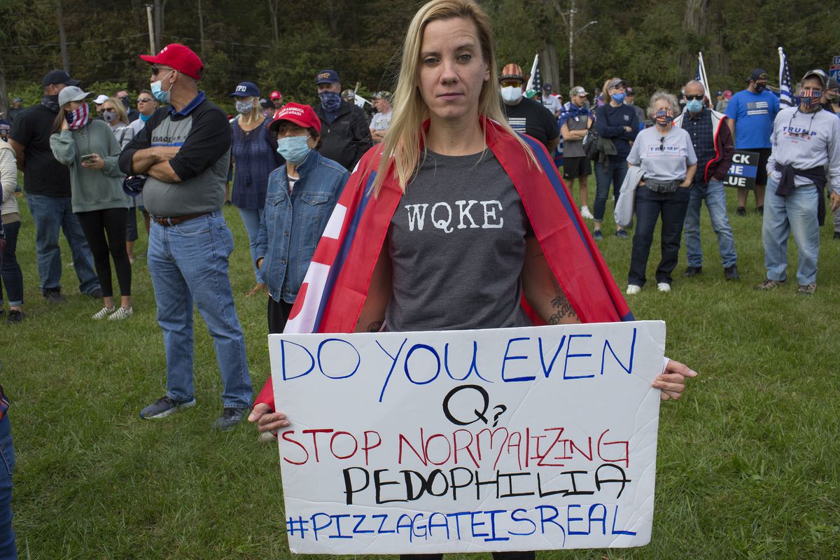 A QAnon supporter wearing a shirt that reads “WQKE” and carrying a sign that reads, “Do you even Q? Stop normalizing pedophilia #pizzagateisreal.”