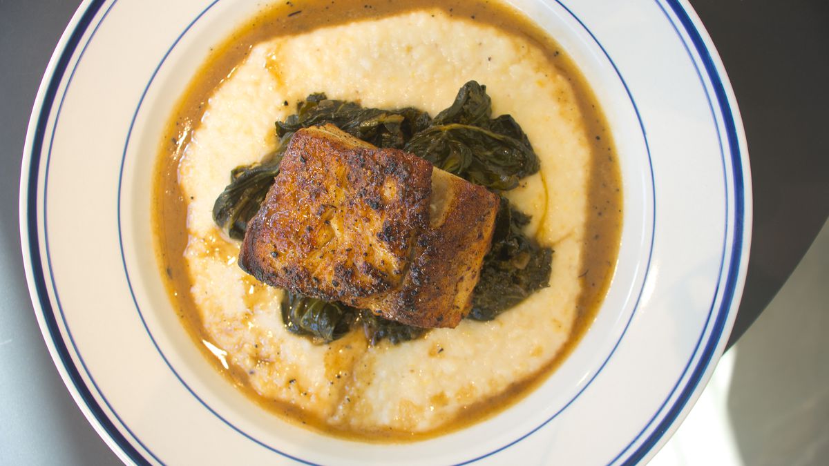 A plate of creamy grits in a brown sauce topped with cooked greens and a piece of brownish fish.