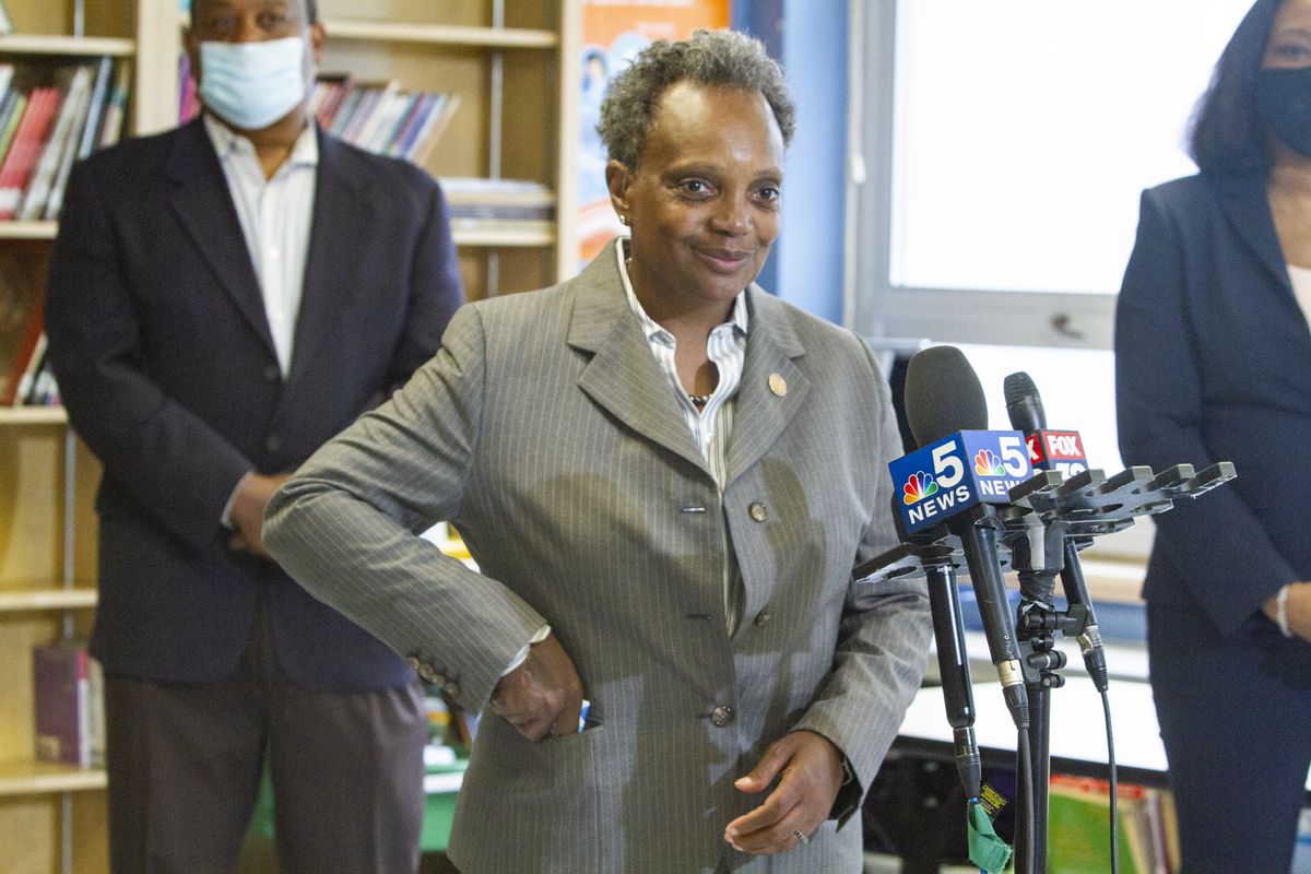 Mayor Lori Lightfoot standing by microphones placing her mask into her jacket pocket.