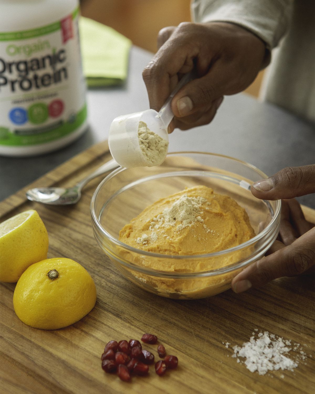 A person mixes protein powder into an orange dip with lemons and pomegranate seeds on the same cutting board