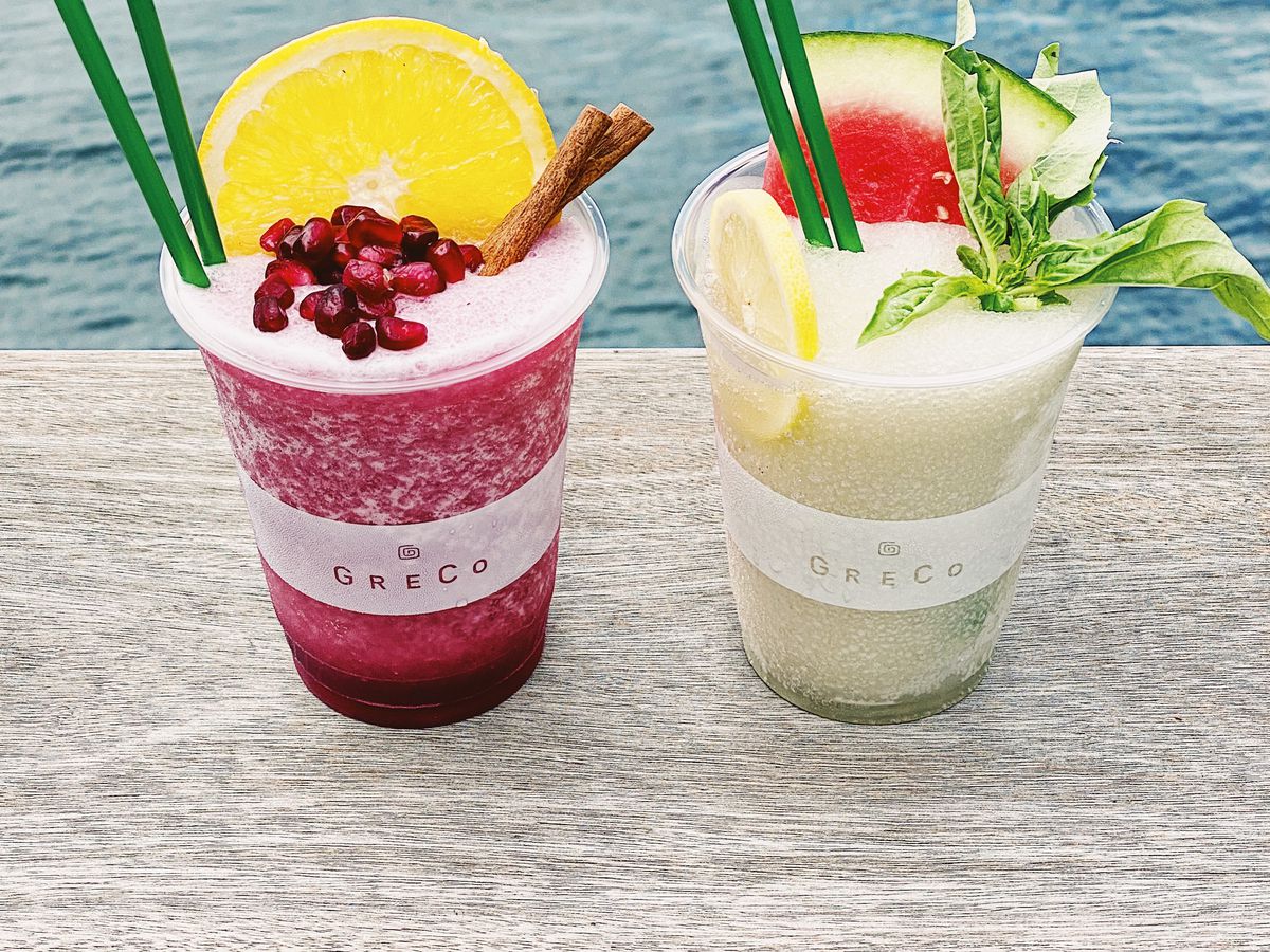 Two plastic cups branded with the restaurant name Greco hold frozen drinks with garnishes and straws. One drink is the color of red wine and the other is pale green.