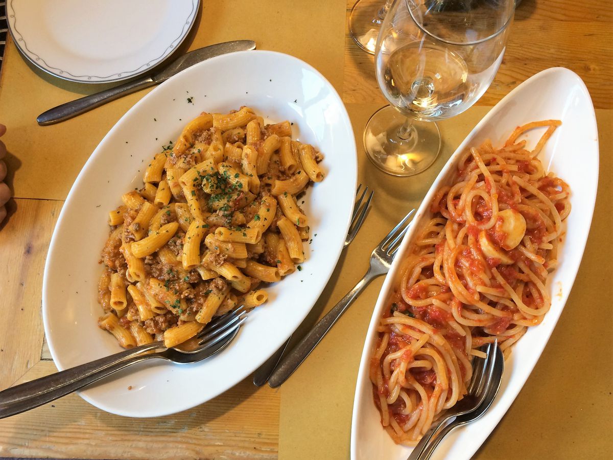 From above, two dishes of pasta in oblong plates beside a glass of wine
