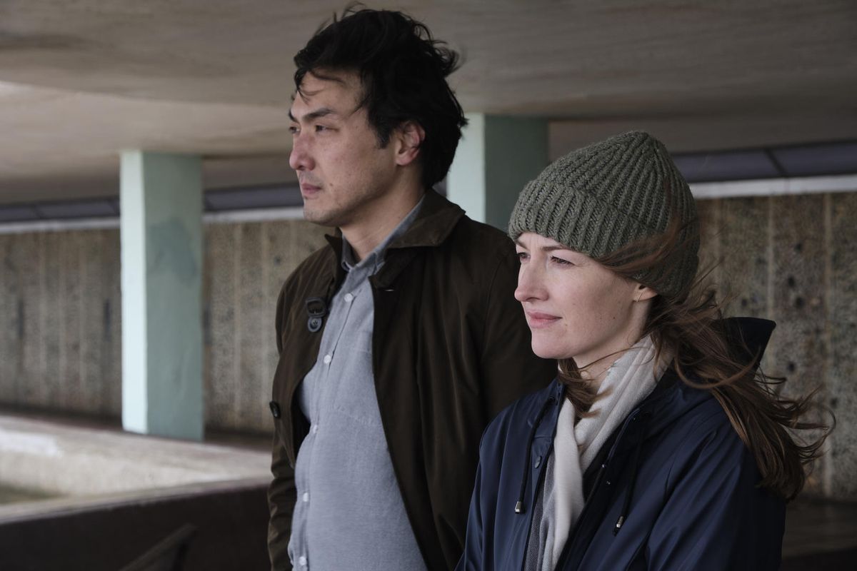 Un hombre que lleva un abrigo marrón (Takehiro Hira) está de pie junto a una mujer con un gorro de lana gris (Kelly Macdonald) y una chaqueta azul, con el pelo alborotado por el viento.