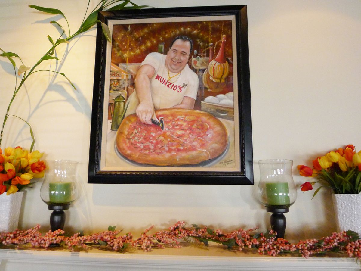 A man enthusiastically cuts a pizza with a wheel in a painting on the mantelpiece flanked by plastic flowers.