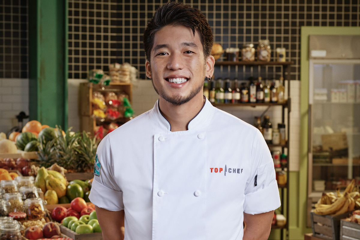 A photo of Shota Nakajima in a “Top Chef” apron with shelves of pantry items in the background