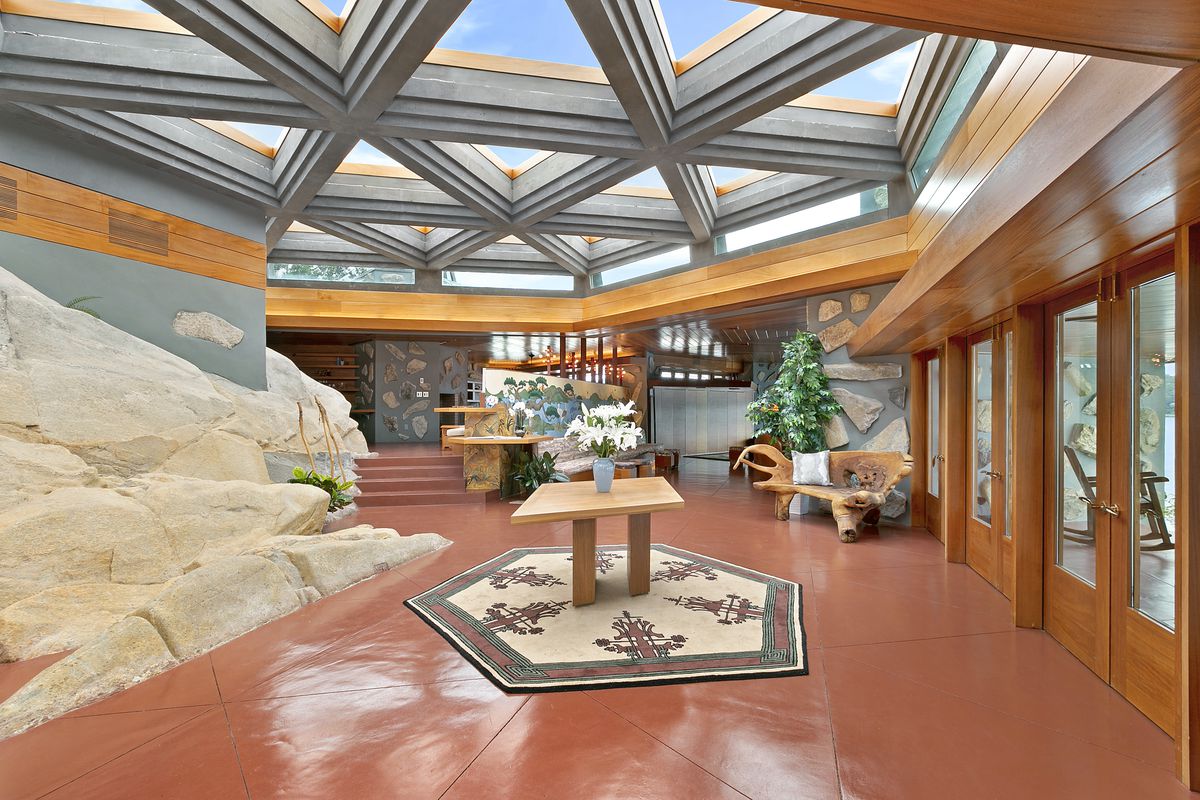 A living area with a ceiling that has partitioned skylights and a red glossy floor. One of the walls has a section that is decorated with rock. There is a table that has a blue vase with white flowers.