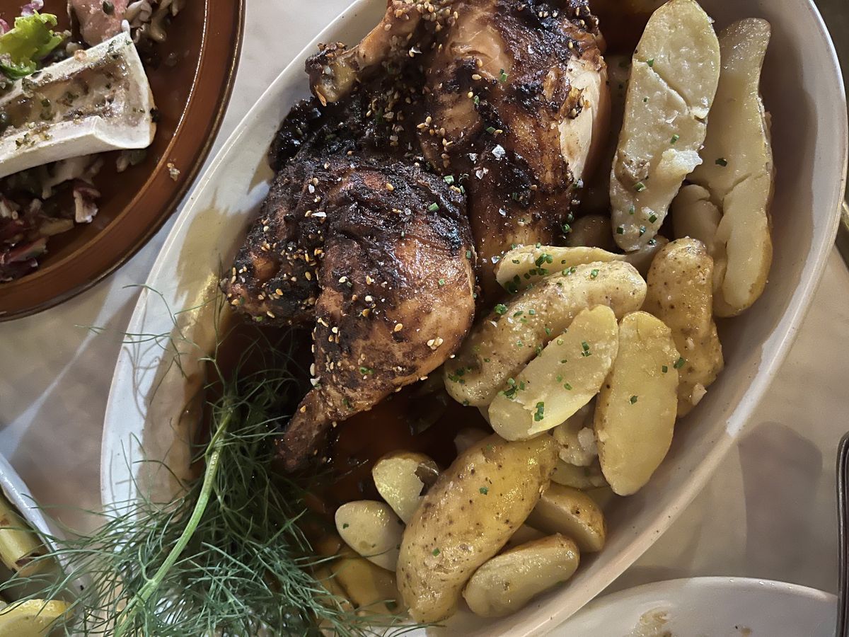 Roasted chicken with a dark skin and a green dill garnish sits in a white plate with fingerling potatoes cut lengthwise and roasted on the side.
